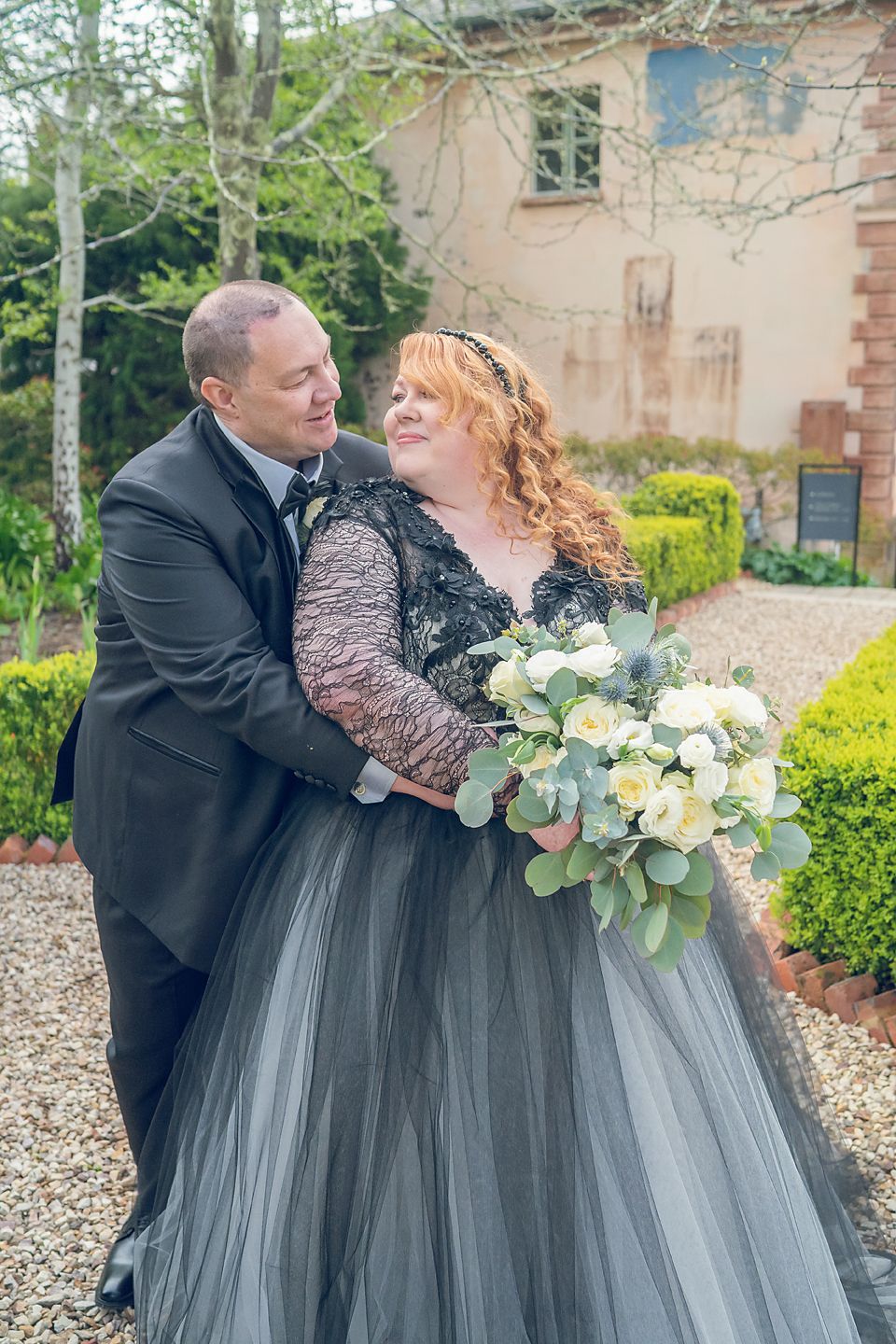 A romantic couple in formal attire sharing tender moments in front of a garden setting with lush greenery.