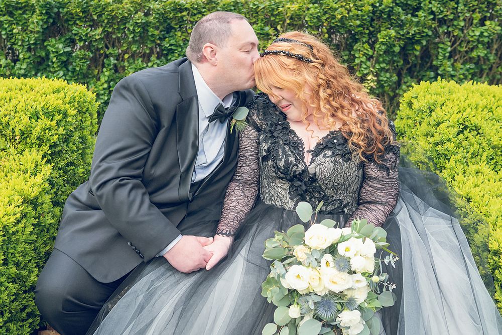 A romantic wedding moment with a couple in black and gray attire holding white flowers against a green hedge backdrop.