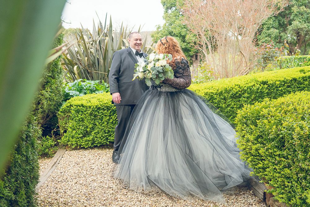 A romantic couple in elegant gray attire poses amongst manicured hedges and garden pathways during a wedding photoshoot.