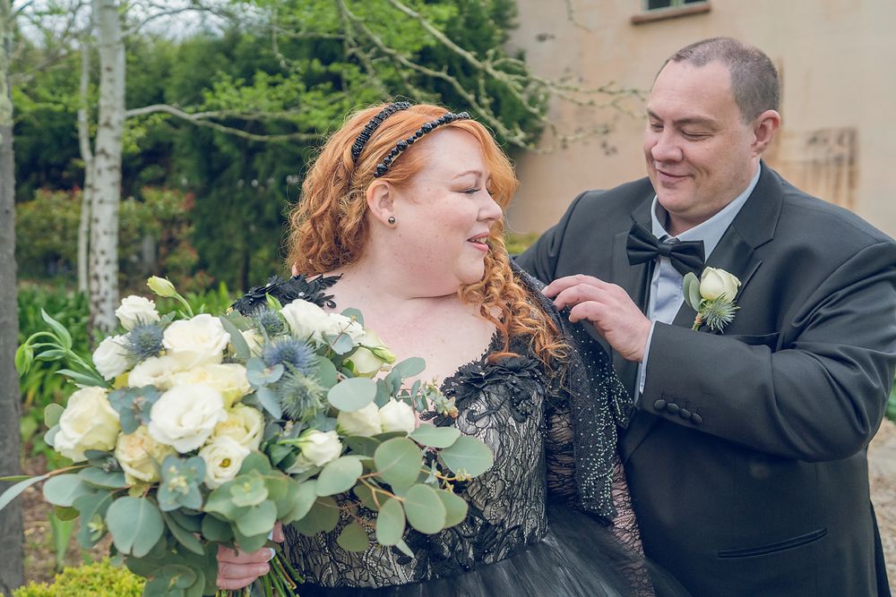 A couple in black formal attire at a wedding celebration with a bouquet of white roses and eucalyptus.