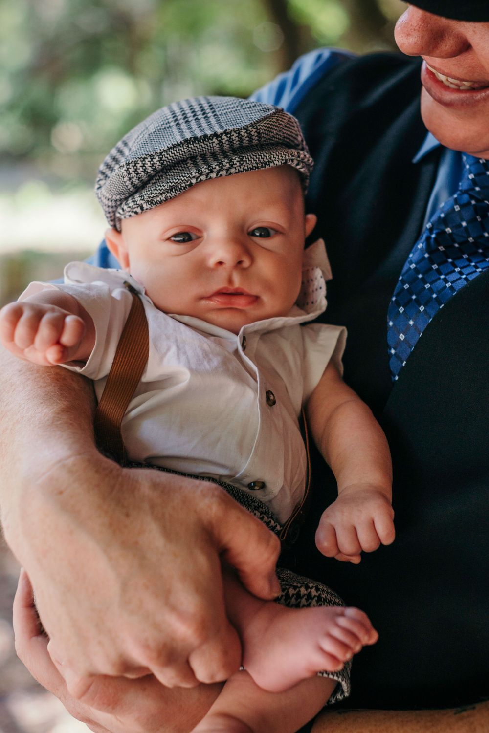 A baby wearing a newsboy cap and white outfit is cradled in someone's arms.