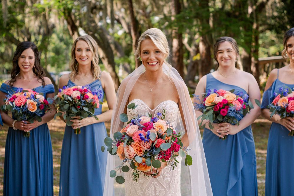 Bridesmaids in blue dresses hold vibrant coral and pink bouquets during outdoor wedding photos.