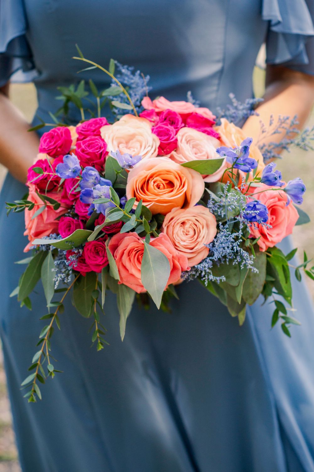 Vibrant bridal bouquet with coral roses, hot pink flowers, and blue delphinium against slate blue fabric.