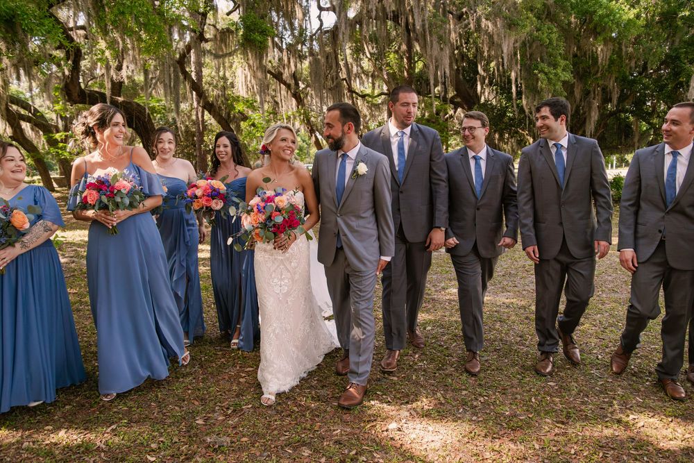 Wedding party in formal attire poses beneath moss-draped oak trees in a scenic outdoor setting.