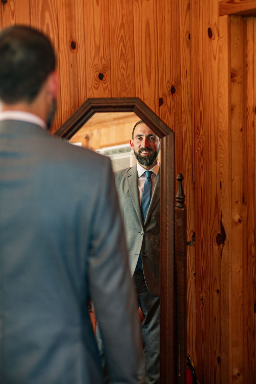 A groom in a grey suit checks his reflection in a rustic wood-paneled mirror before the ceremony.