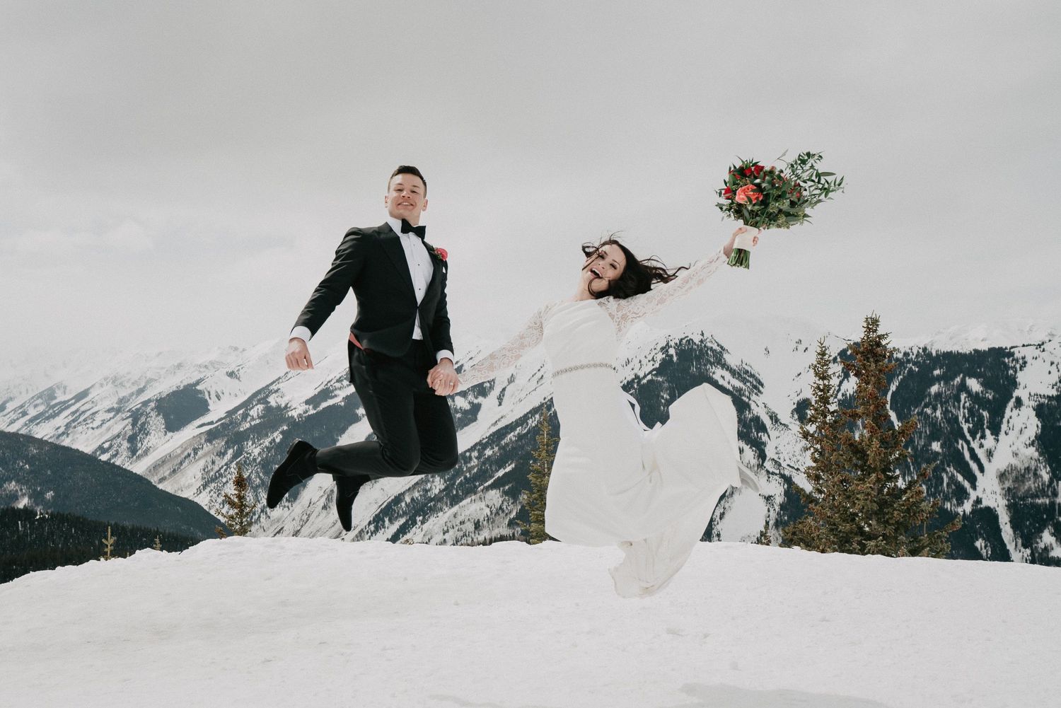A couple in wedding attire leaps joyfully against a backdrop of snow-covered mountains in winter. Elopement photography in Aspen