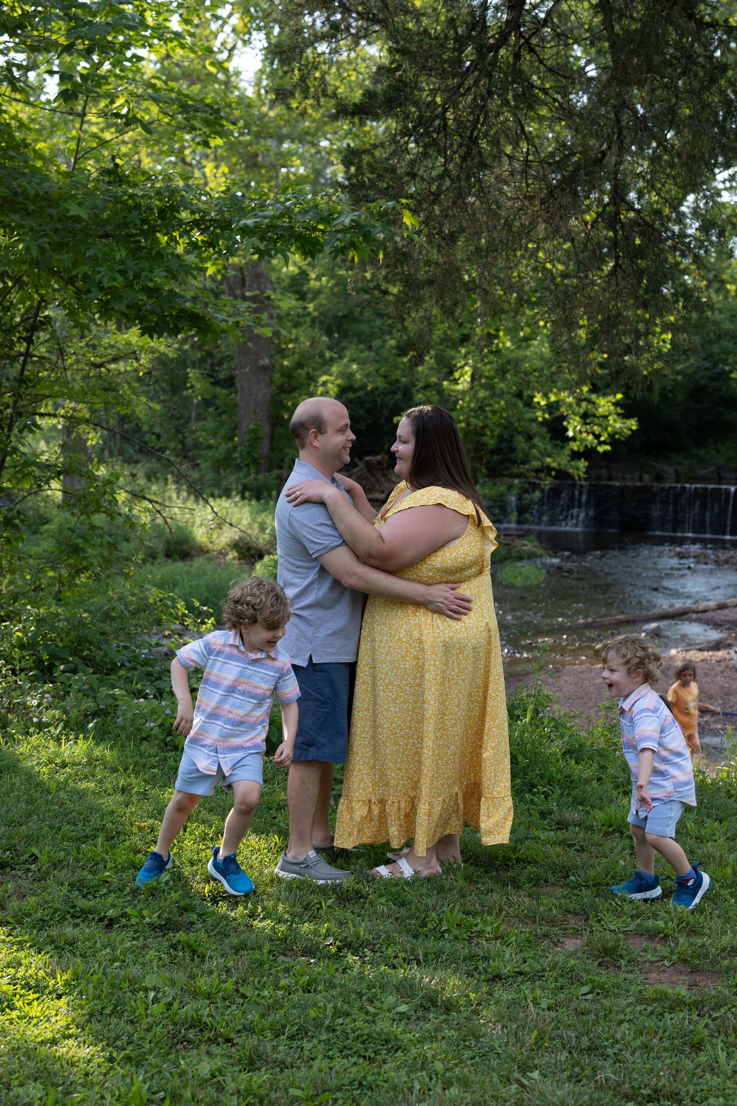 A couple embraces near a waterfall while two children play on the grass in a serene park setting.