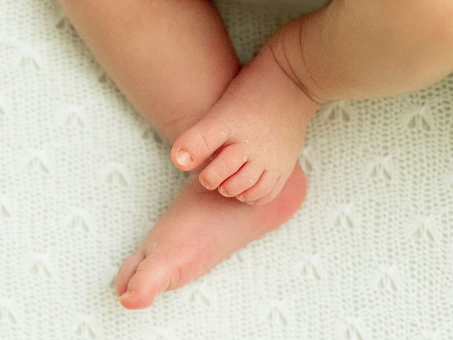 Small newborn flakey baby feet adorably resting on a soft white textured blanket or bedspread.