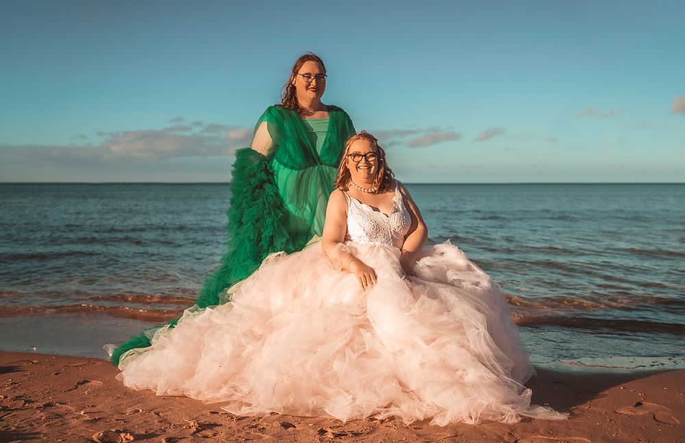 Two people in elegant gowns, one in emerald green and one in blush pink, pose together on a beach at sunset.
