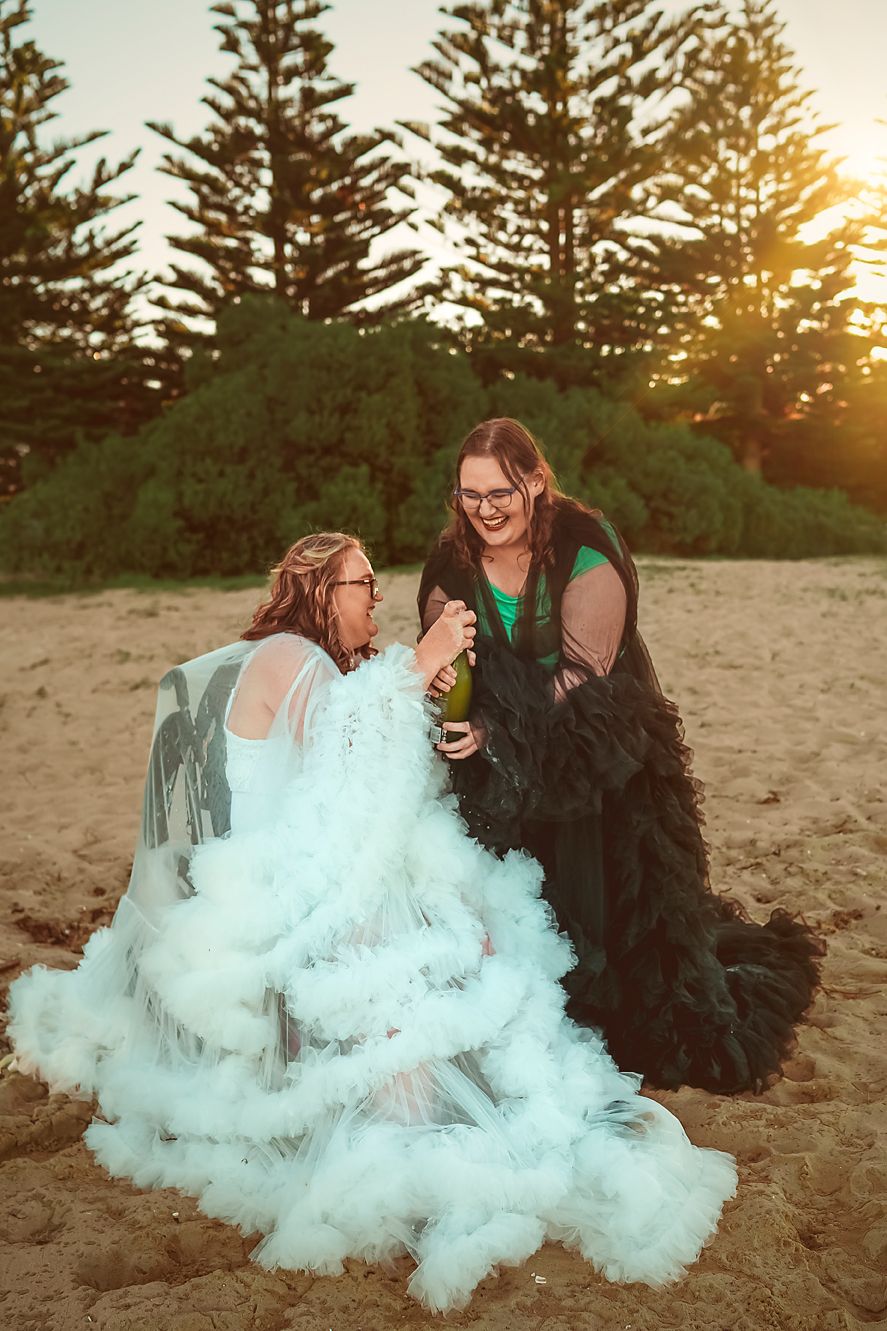 Two people sitting on beach sand surrounded by teal-colored smoke during sunset with evergreen trees in the background.