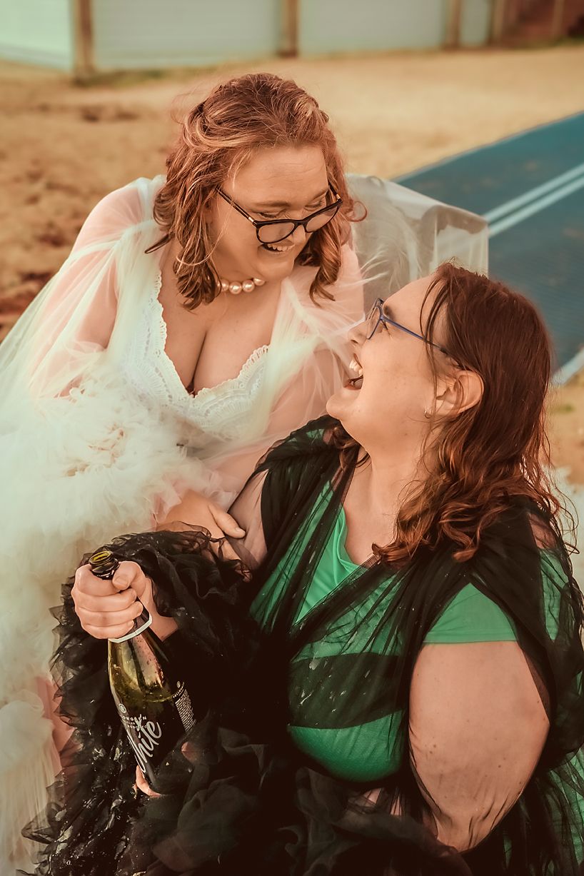 Two women in wedding attire share a joyful moment together while holding champagne and laughing.