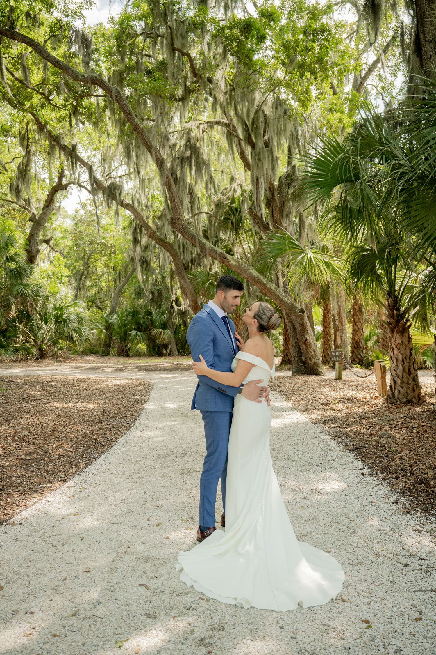 A couple embraces on a shell path beneath Spanish moss and palm trees at a romantic Southern garden venue.