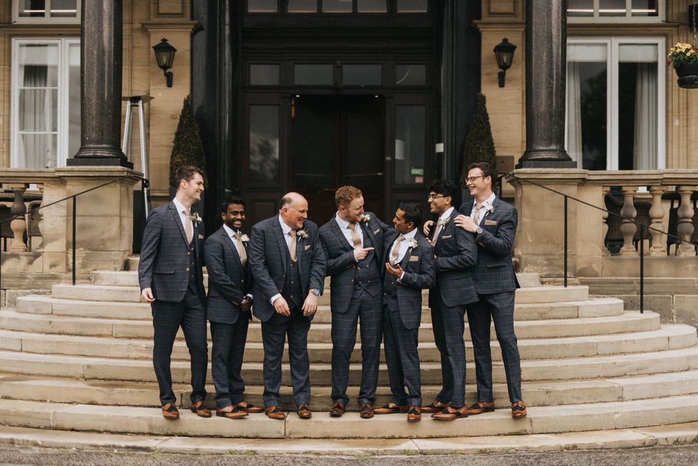 A wedding party in navy blue suits stands laughing together on grand stone steps of an elegant historic building.
