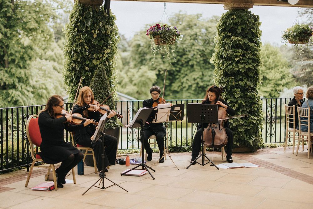 String quartet musicians perform on outdoor patio with elegant ivy-covered pillars and garden backdrop.