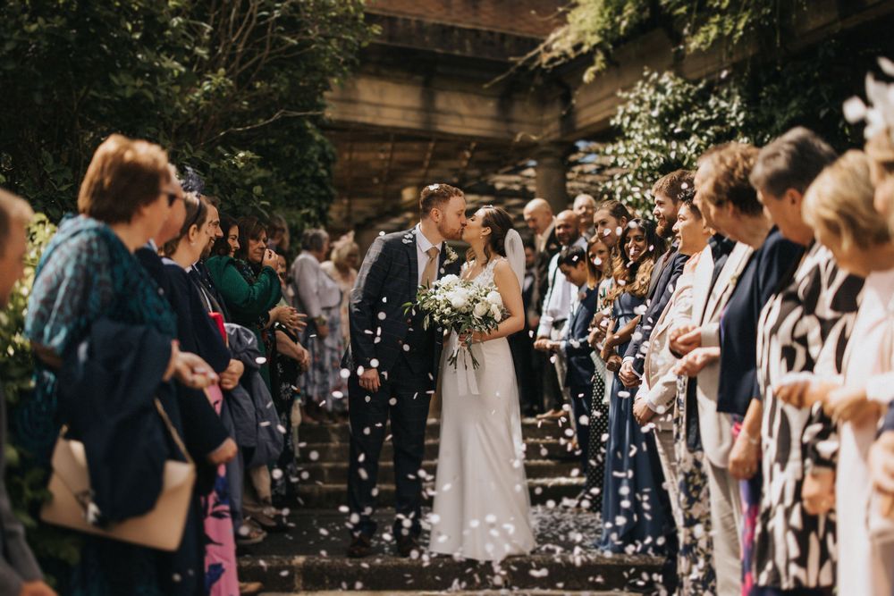 Confetti falls over wedding guests lining both sides of a path as they celebrate at an outdoor ceremony.