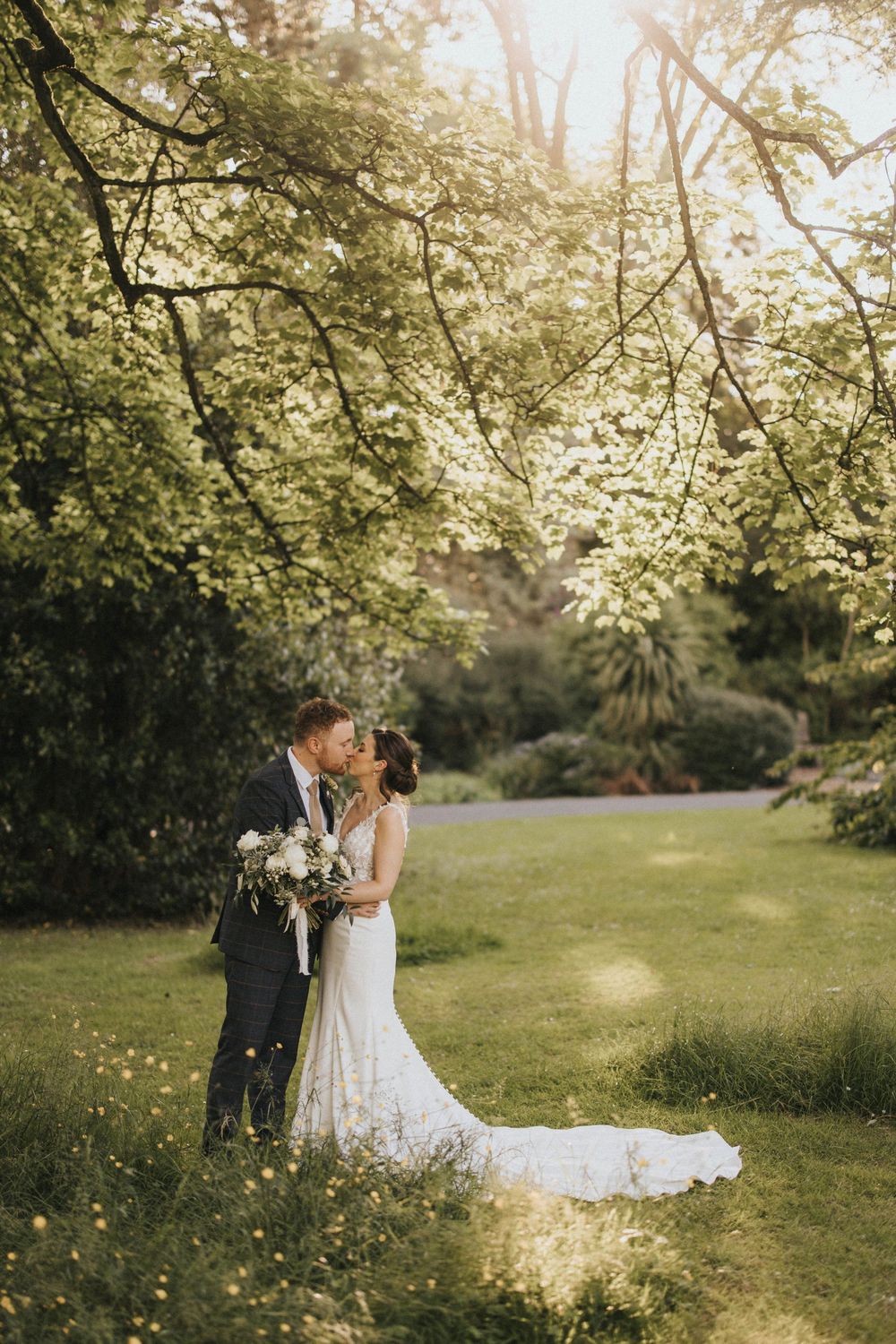 A romantic wedding couple stands beneath blooming cherry blossom trees in a sun-dappled spring garden setting.