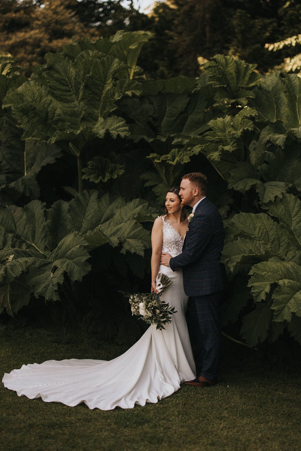 A couple in wedding attire poses against a backdrop of giant tropical leaves in a dramatic garden setting.