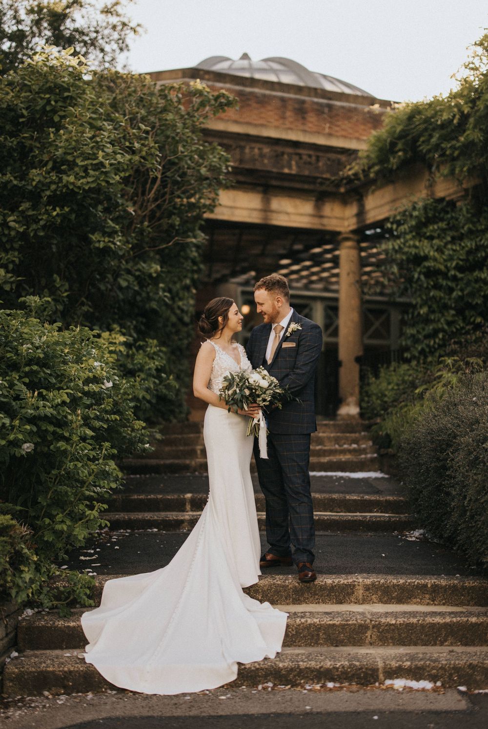 A romantic wedding couple stands on stone steps surrounded by lush greenery and architectural pillars at sunset.