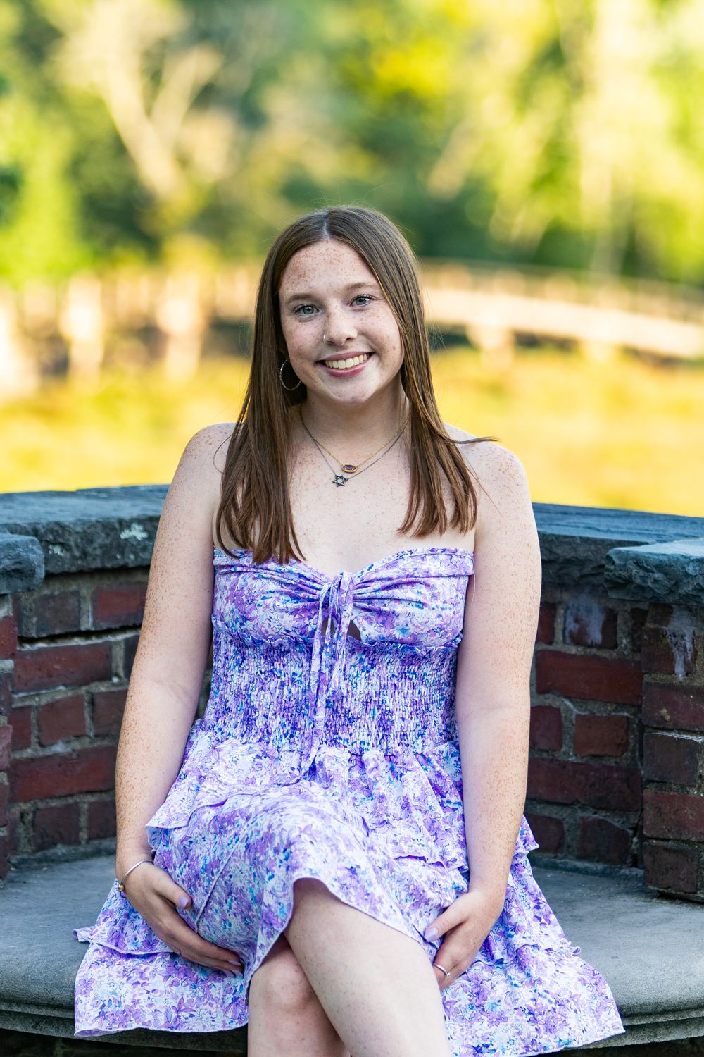 A person in a purple floral summer dress sits on a brick wall on a sunny day with blurred greenery in background.