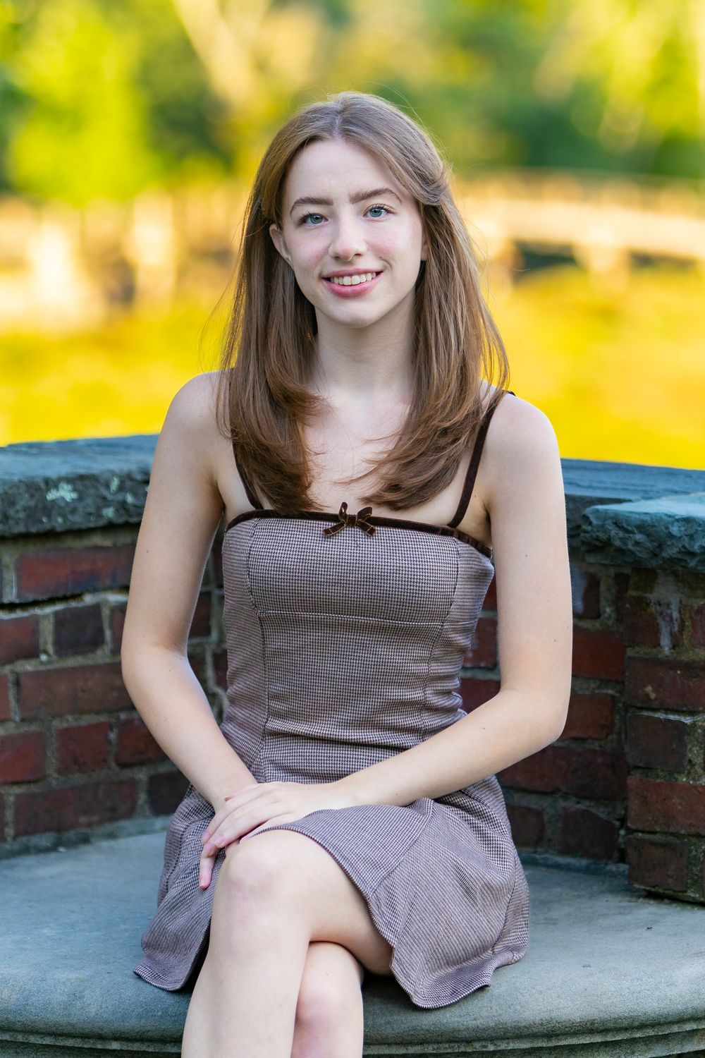 A young woman in a brown dress sits on a stone bench in a garden with bright green foliage in the background.