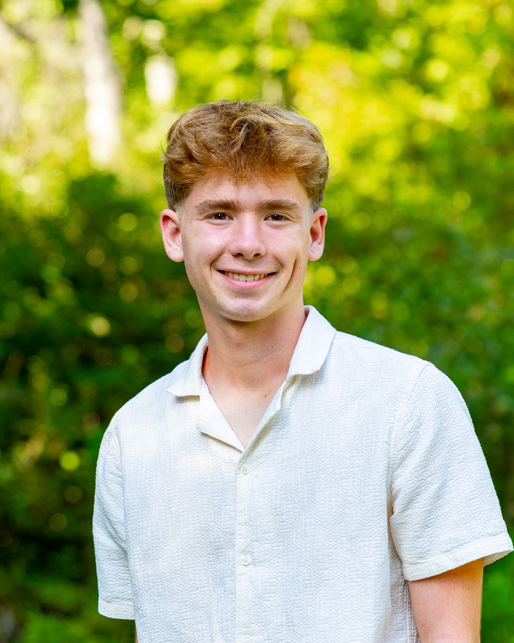 A person wearing a white polo shirt smiles while standing in front of vibrant green foliage outdoors.