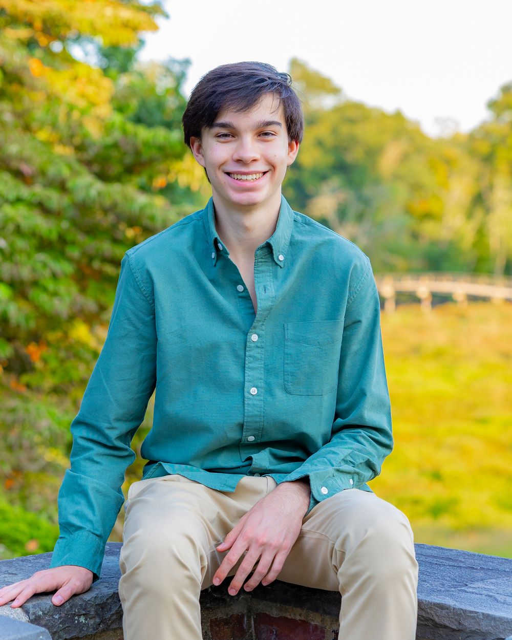 A person in a teal button-down shirt sits outdoors on a sunny autumn day with yellow foliage in the background.