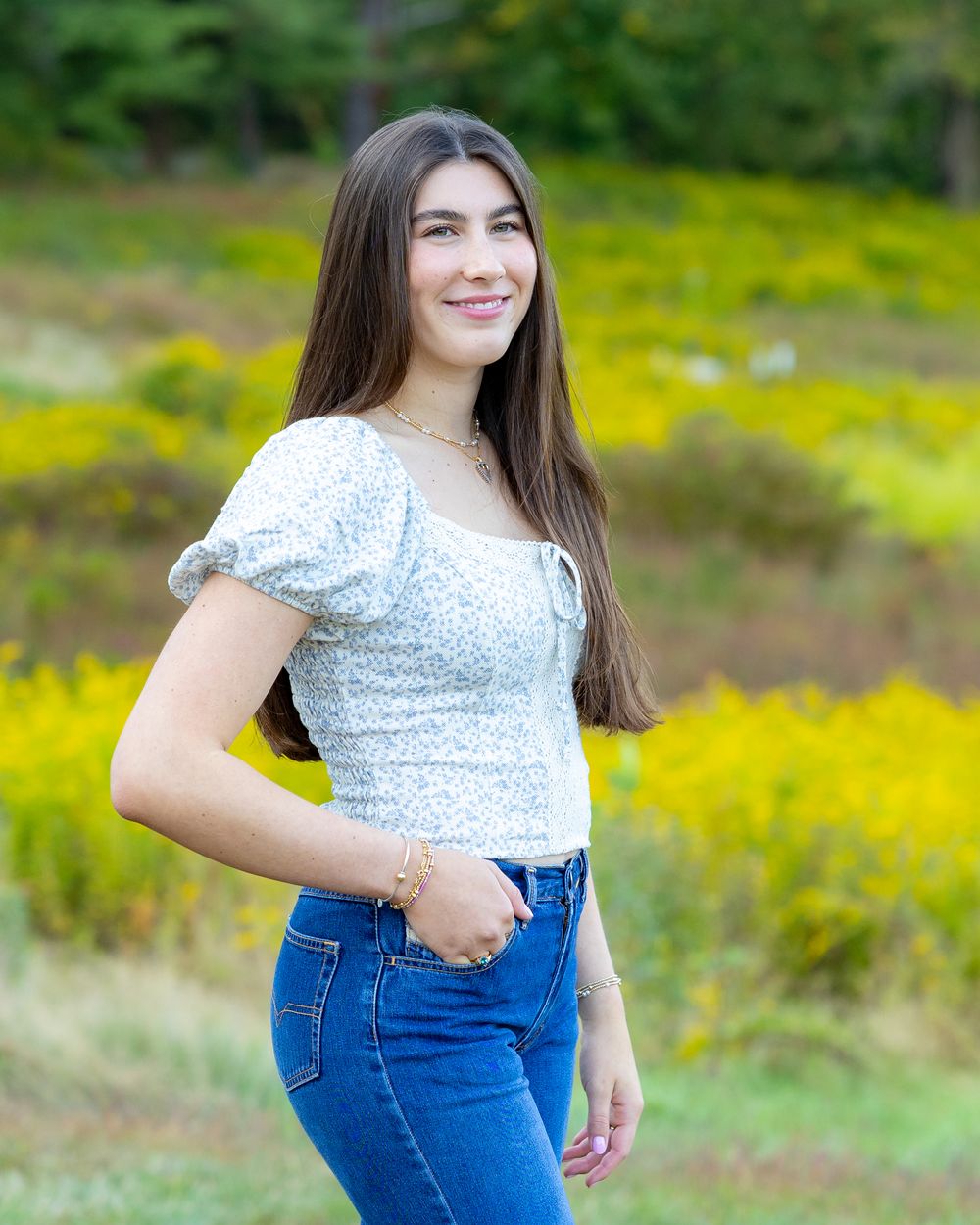 A person in a floral puff sleeve top and blue jeans poses in a field of yellow wildflowers on a sunny day.