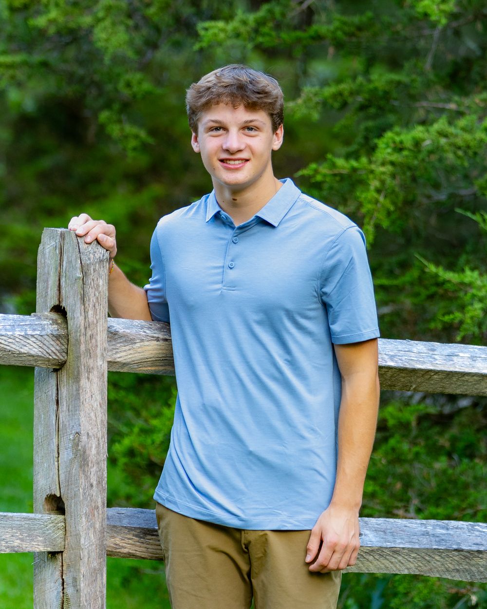 Person in a light blue polo shirt stands by a rustic wooden fence with green foliage in the background.