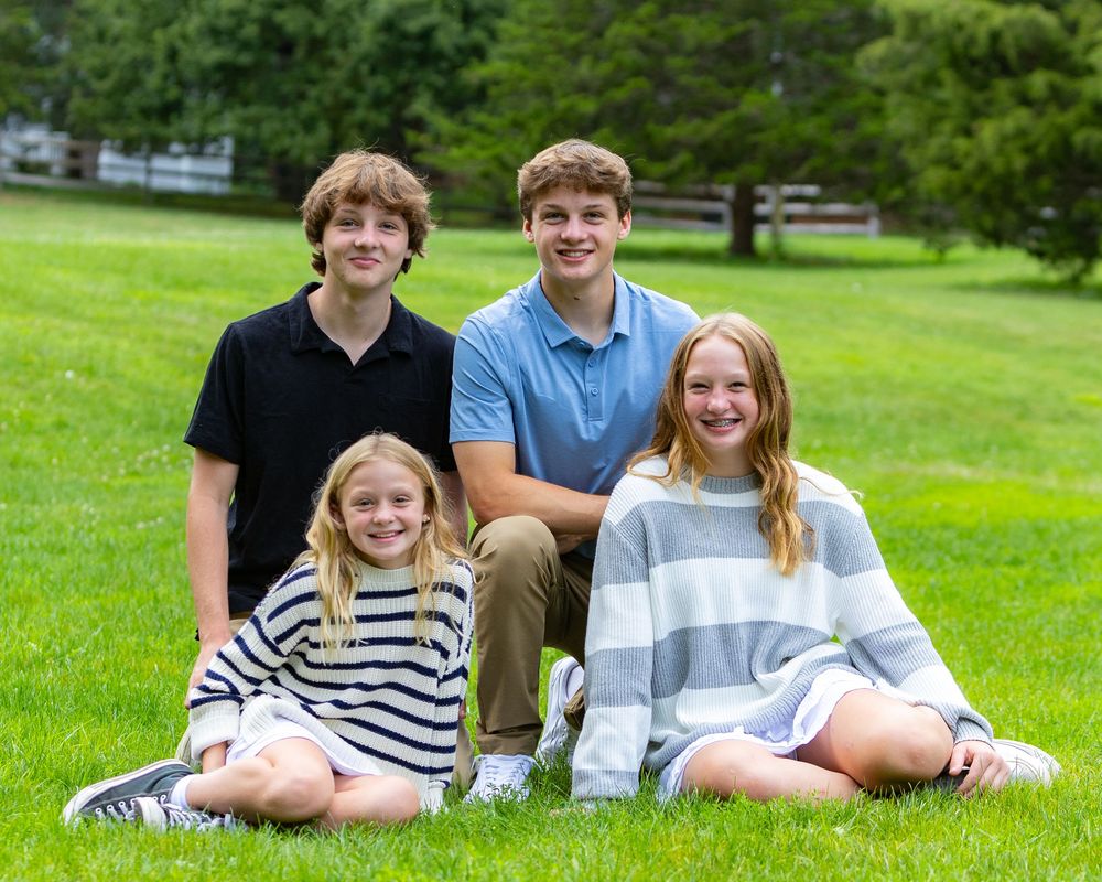 Four young people sitting together on grass in a park with trees and white fence in background.