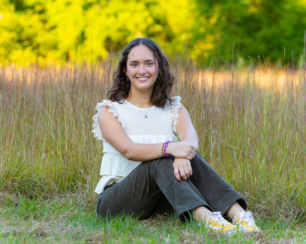 A young woman with dark wavy hair sits in a grassy field wearing a white top and dark pants during golden hour.