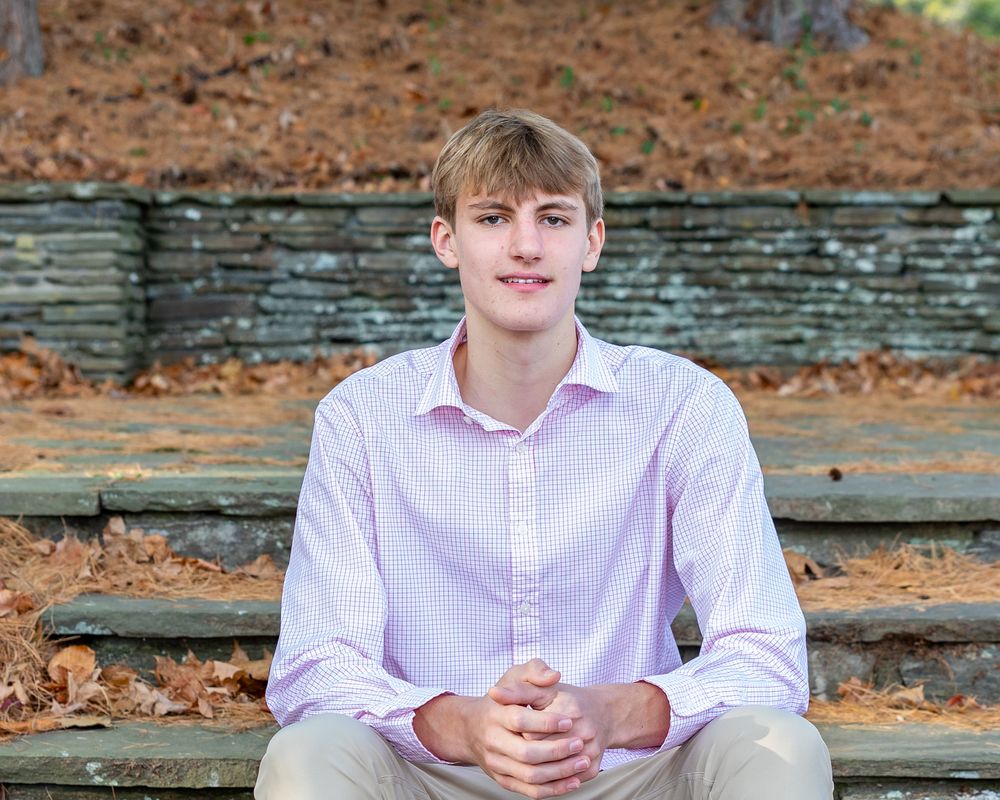 A young man with a pink dress shirt sits on a rustic wooden bench surrounded by fallen autumn leaves.