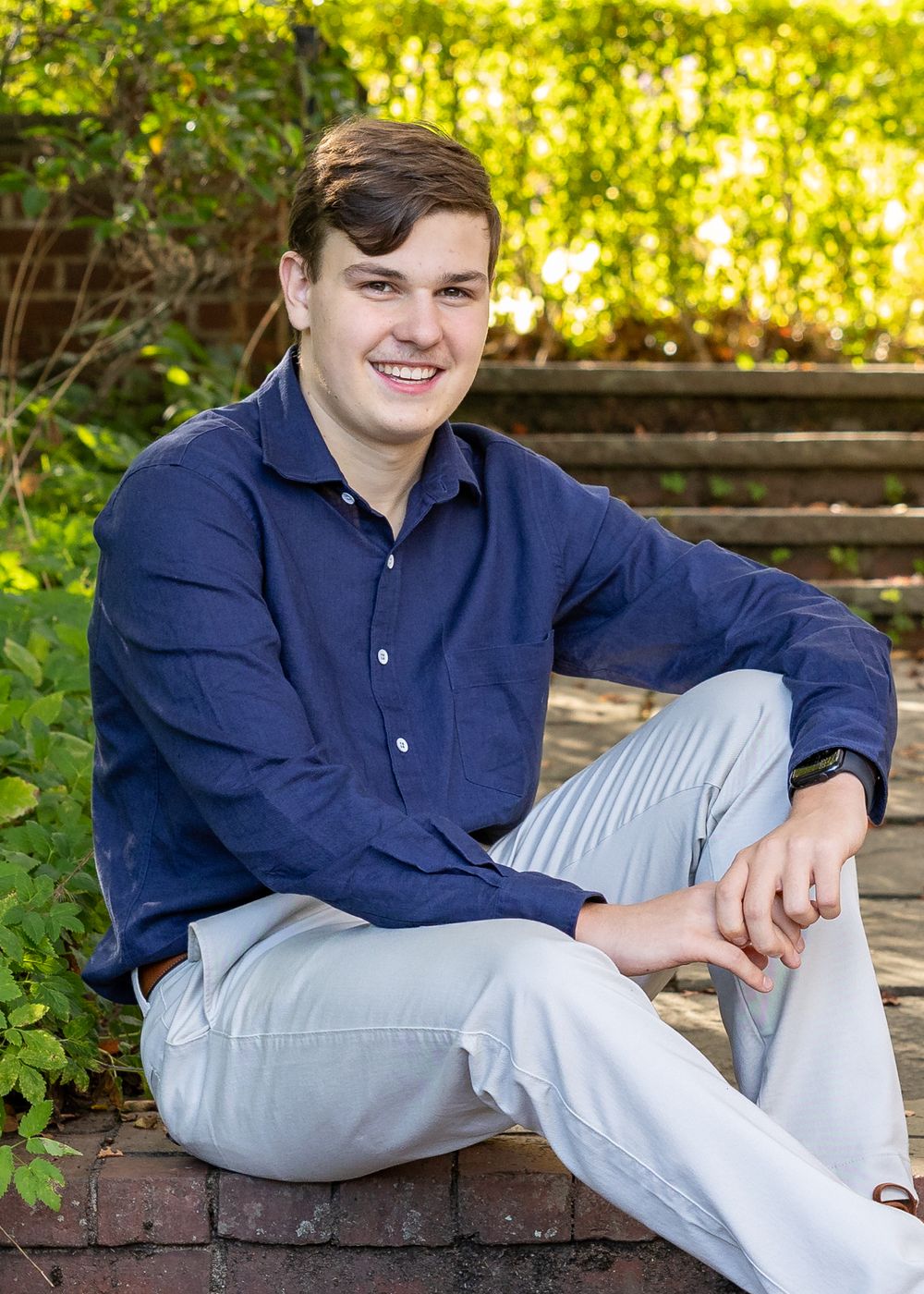 A person in a navy button-down shirt and light gray pants relaxes casually on stacked brick pavers in a sunny garden setting.
