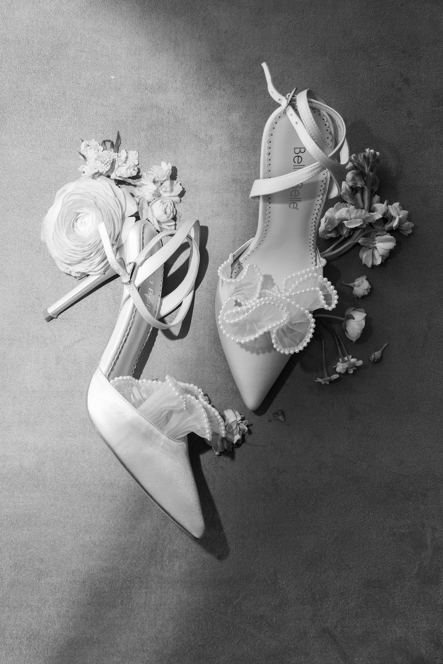 Elegant white bridal shoes with pearl details and delicate floral decorations arranged on grey surface in black and white.