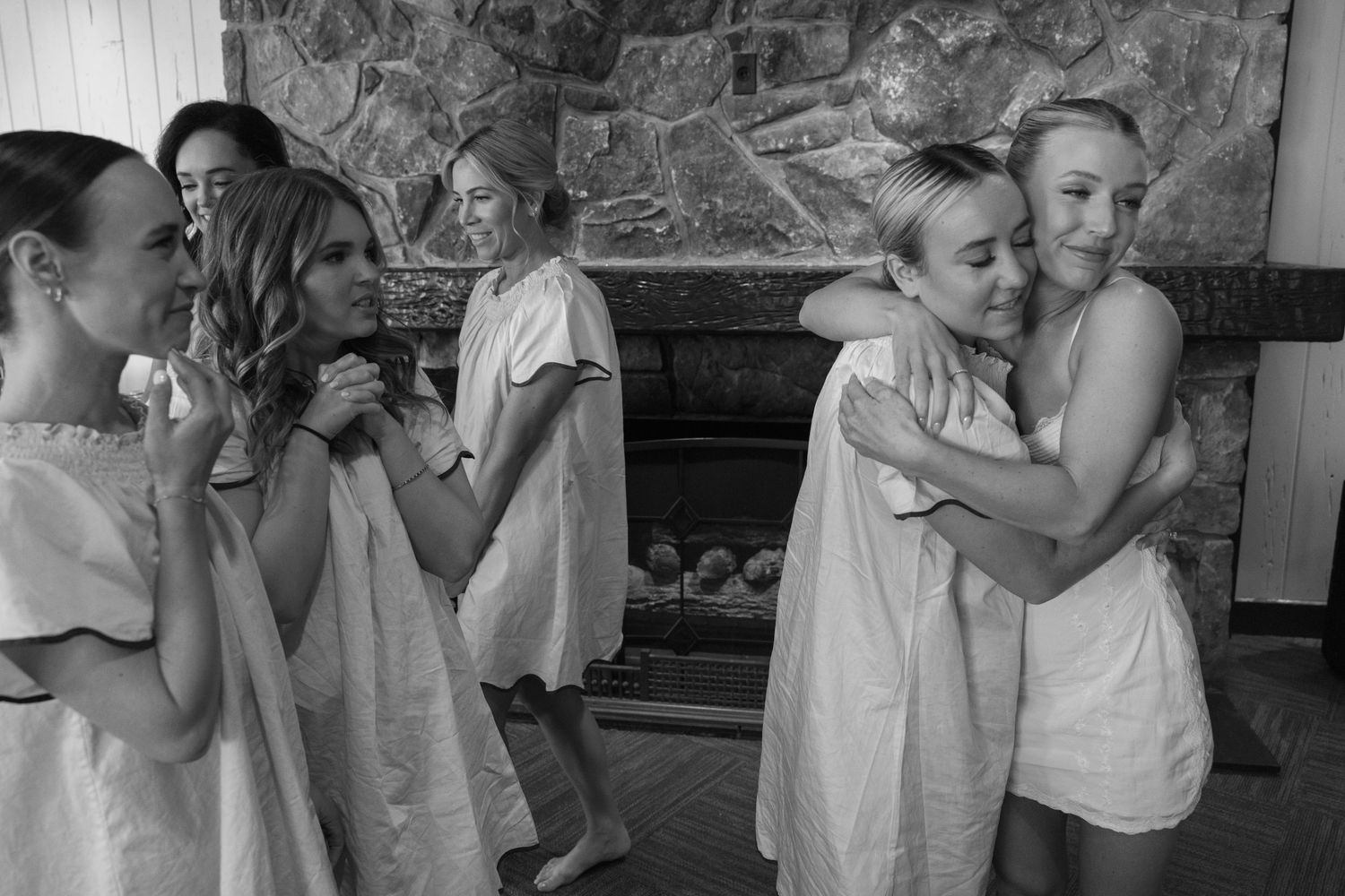 Black and white photograph of people in light colored garments embracing in a rustic interior space with stone walls.