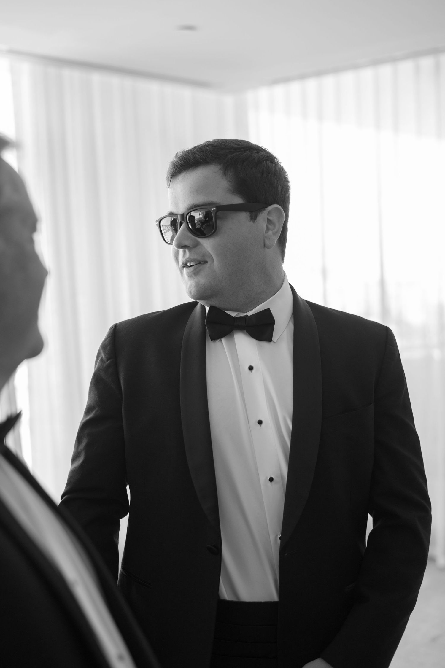 A person in a formal black tuxedo and bow tie wearing sunglasses smiles while engaging in conversation.