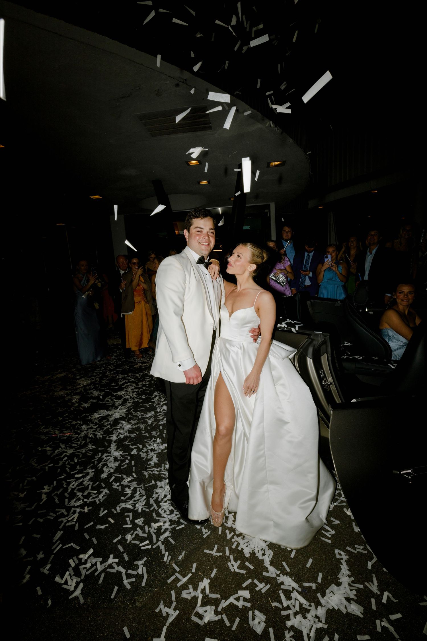 Two people in white formal attire celebrate at their wedding reception with confetti falling around them on a dark dance floor.