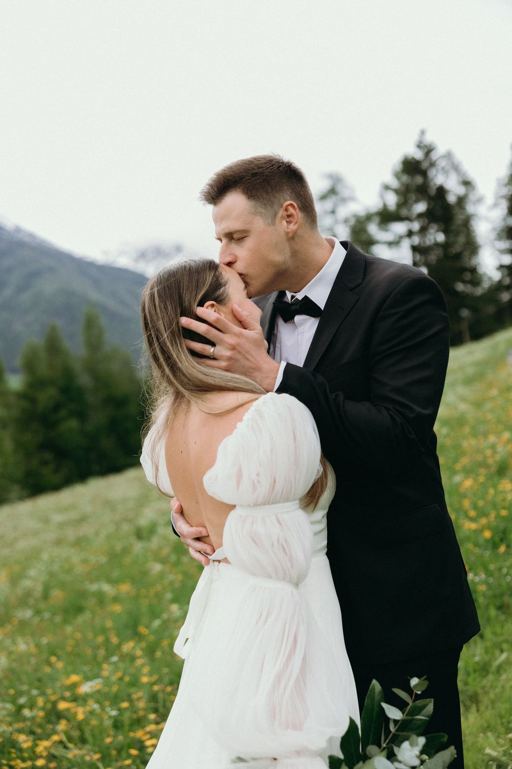 A couple in wedding attire share a romantic kiss on a mountainside meadow filled with wildflowers.