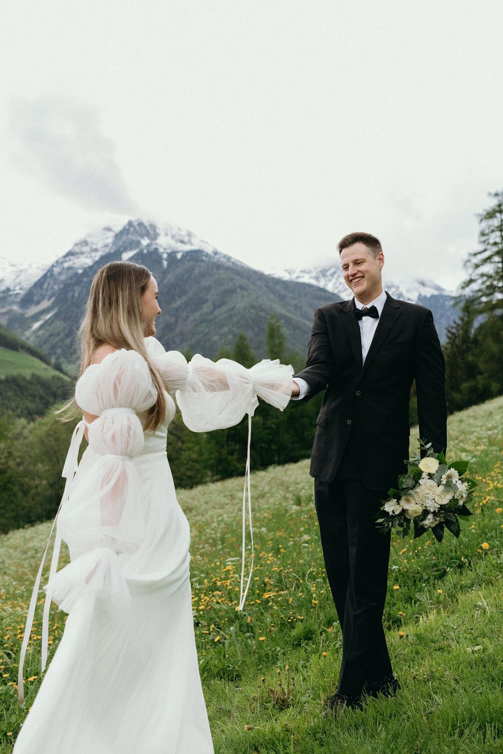Newlyweds share a joyful moment on a hillside with snow-capped mountains in the background.