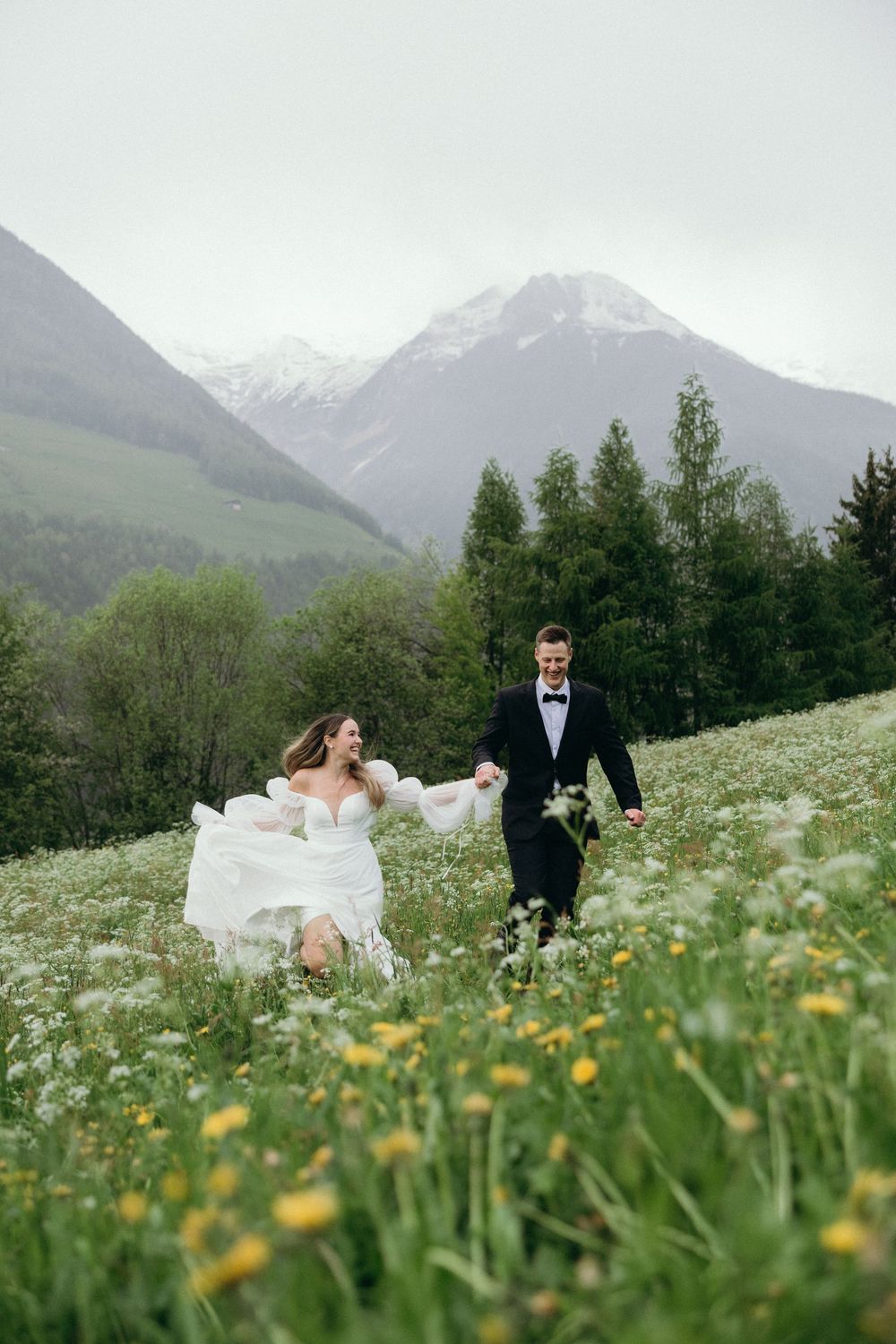 A couple in wedding attire stand in a wildflower meadow with snow-capped mountains in the background.