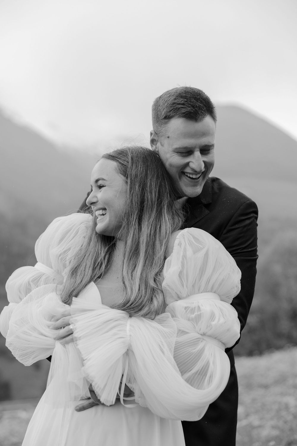 A candid black and white photo captures newlyweds laughing together against a mountain backdrop.