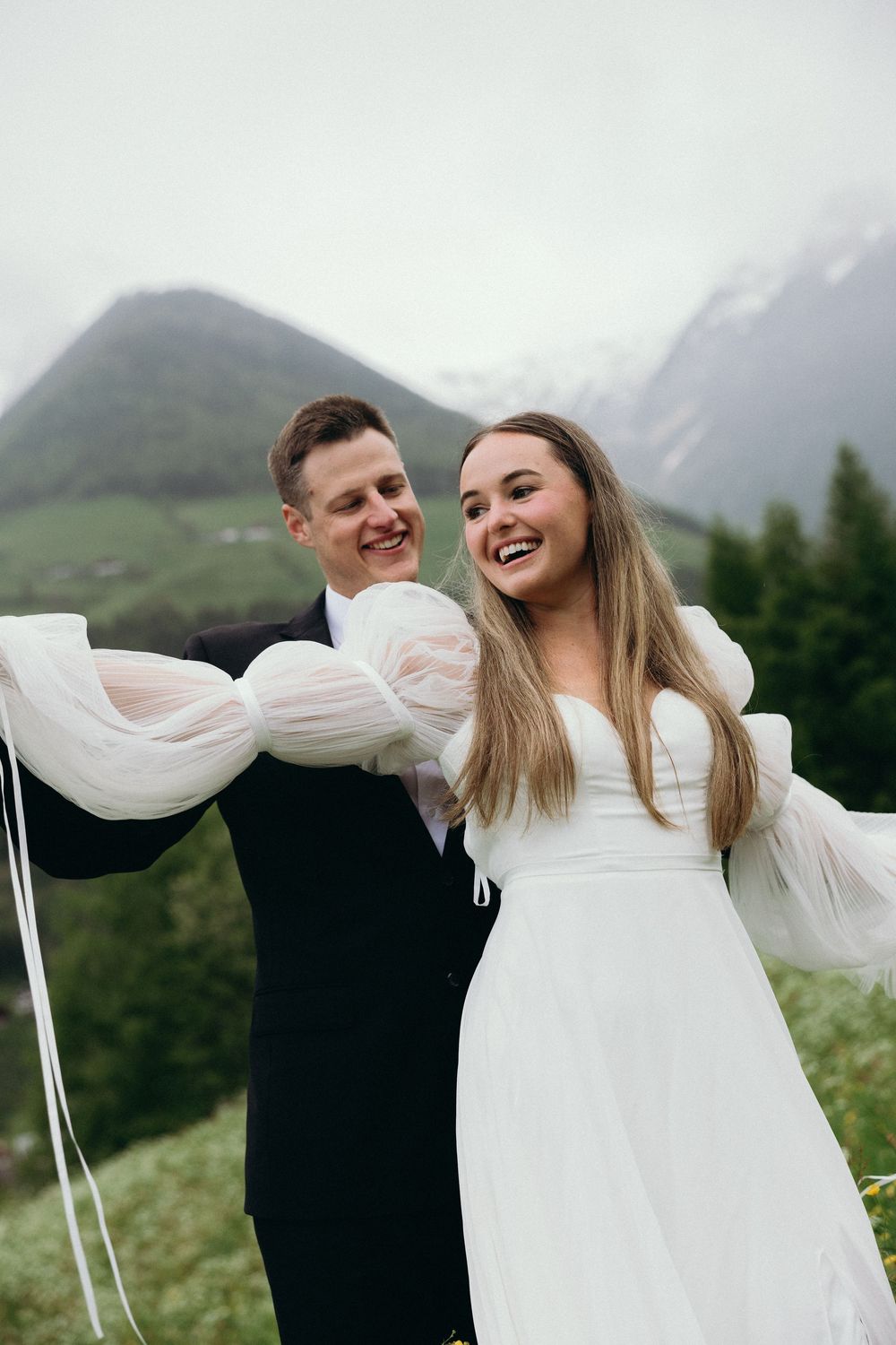 Romantic wedding couple embraces against a mountain backdrop with flowing white dress and dramatic cloudy skies.