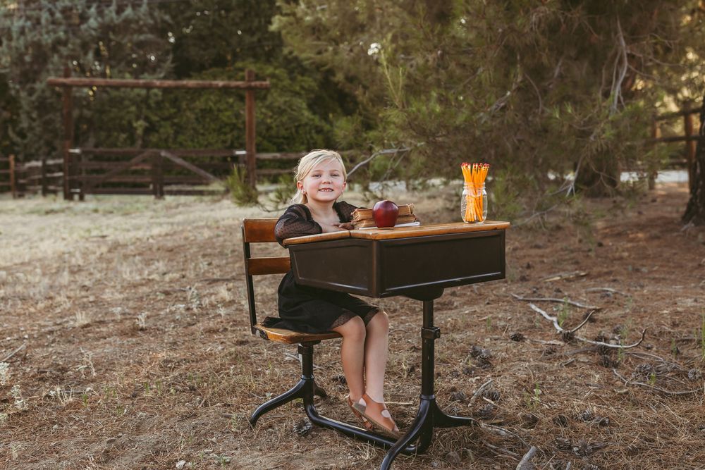 A child sitting at an antique wooden school desk outdoors with fall foliage in the background.