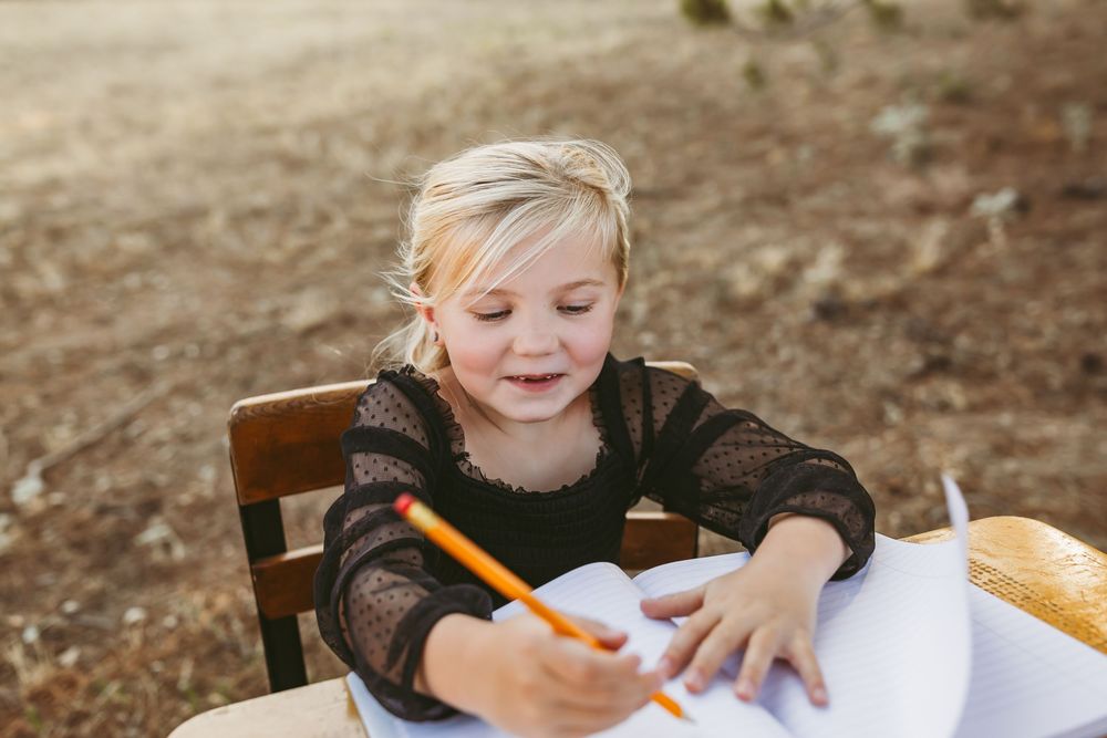 Young child writing with a pencil at a wooden table outdoors in natural sunlight.