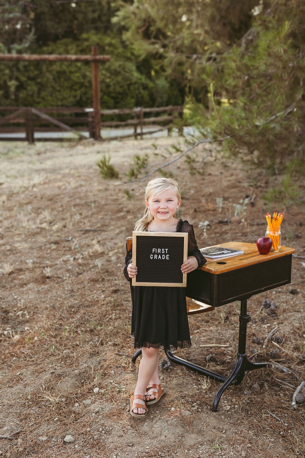 Small child stands by vintage school desk outdoors holding black letter board with supplies on desktop surface.