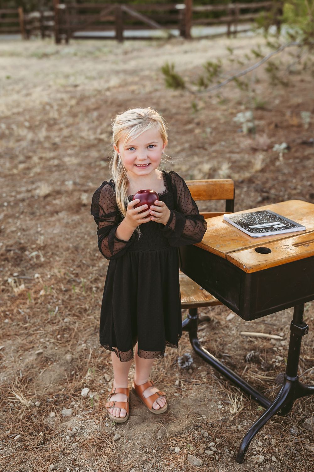 A child in black dress holding a drink stands beside an antique school desk outdoors in a dirt area.