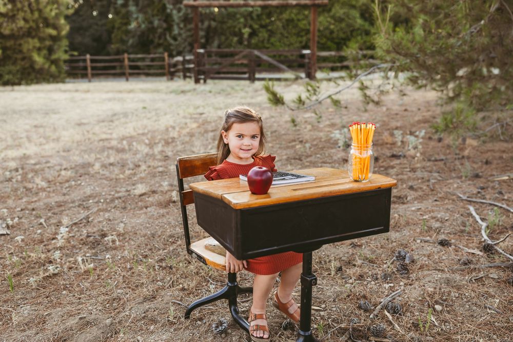 Small wooden student desk sits outdoors in nature with a drink and snack on top while someone sits at it.