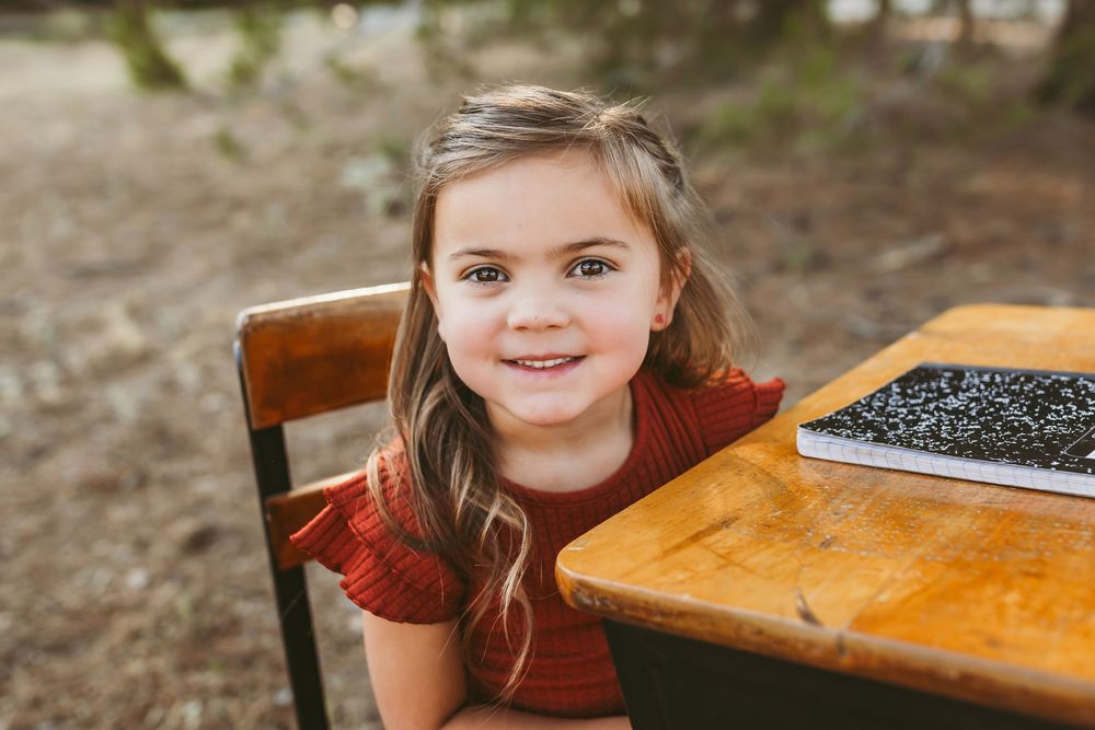 Young student in a red shirt sits at a wooden desk with a composition notebook outdoors.