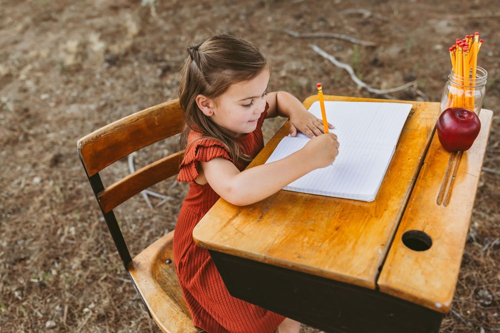 Student in a red dress studiously writing at an old wooden desk with pencils in glass jar outdoors.