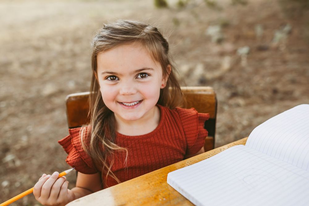 A child in a red turtleneck sweater sits at a desk while looking at the camera and grinning.