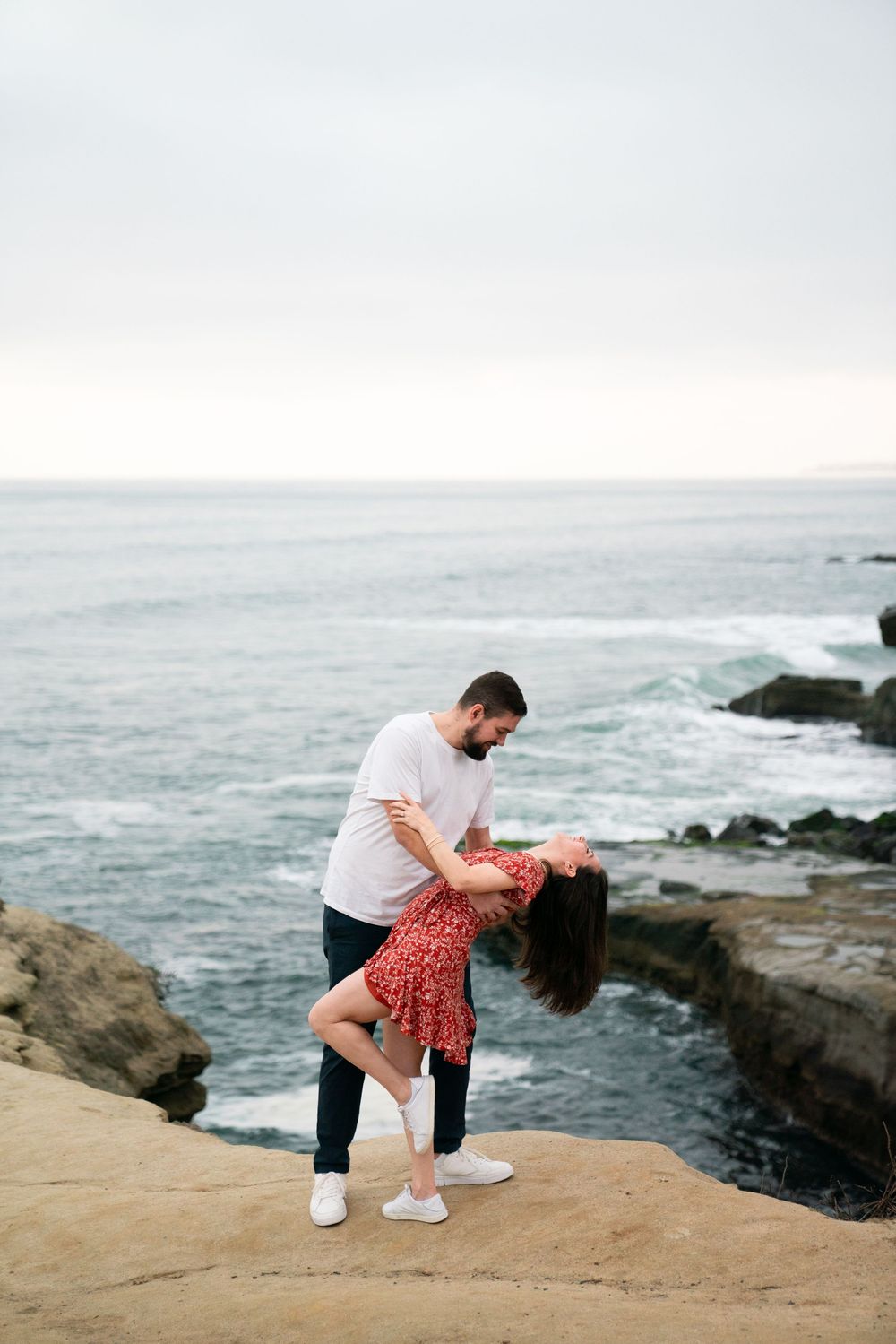 A couple shares a romantic moment on a rocky coastal cliff overlooking the ocean as waves crash below.